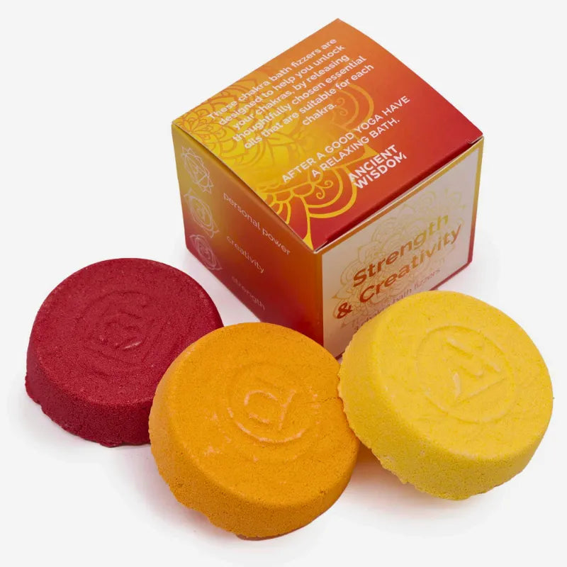 Trois bombes de bain moussantes (jaune, orange, rouge) avec motifs gravés pour les chakras, accompagnées d’une boîte cadeau "Strength & Creativity" au design vibrant. 